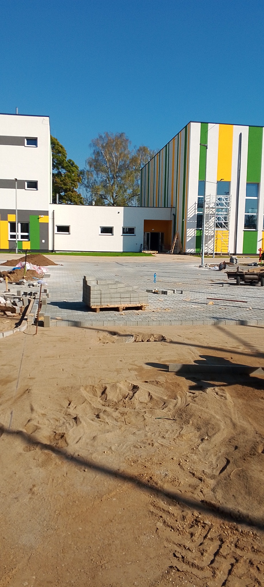 Plac budowy z częściowo ułożoną szarą kostką brukową, paleta z kostką w tle, nowoczesny budynek z elementami w kolorach żółtym i zielonym.