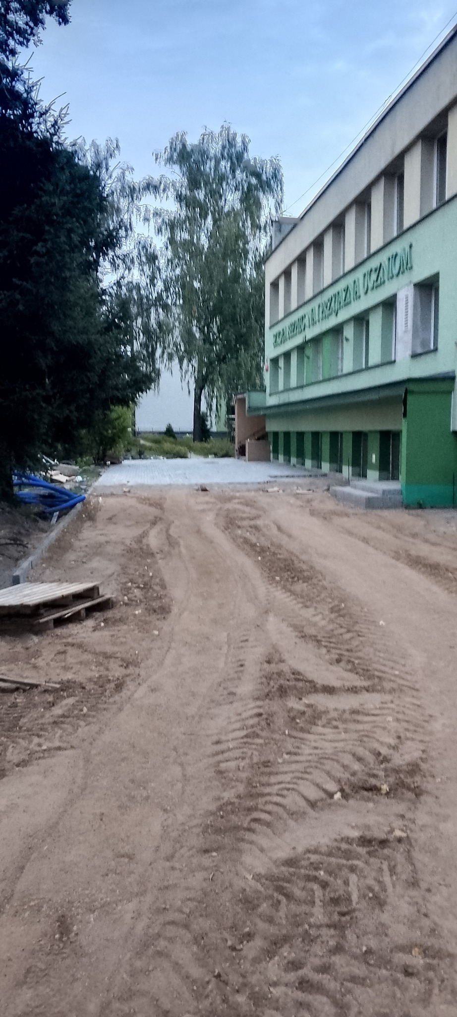 Nowa nawierzchnia z kostki brukowej przed budynkiem, widoczne ślady opon i paleta drewna, w tle drzewa i fragment budynku z napisem.