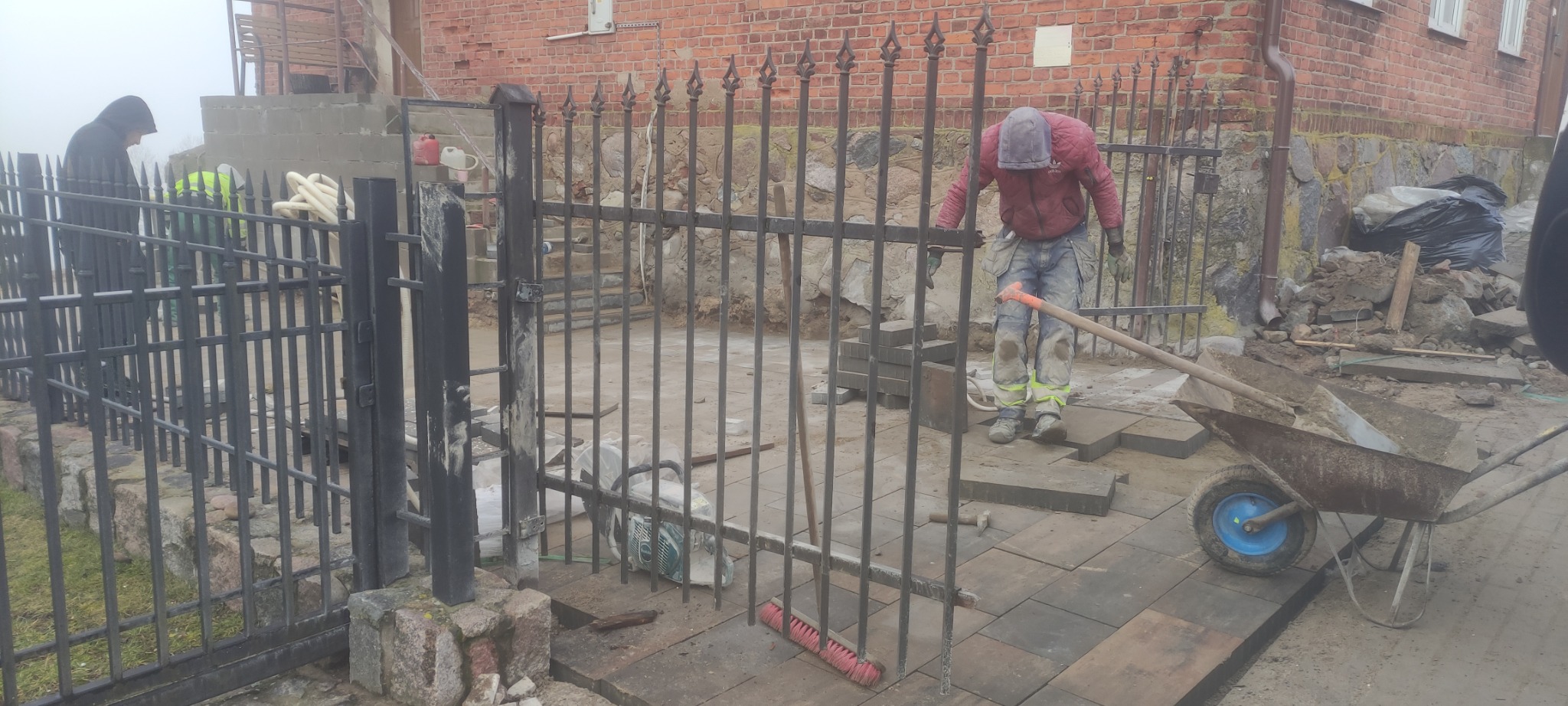 Prace brukarskie w toku: robotnicy układają szare kostki obok metalowego ogrodzenia, widoczny wózek z niebieskim kołem, piła do cięcia kostki i narzędzia brukarskie.