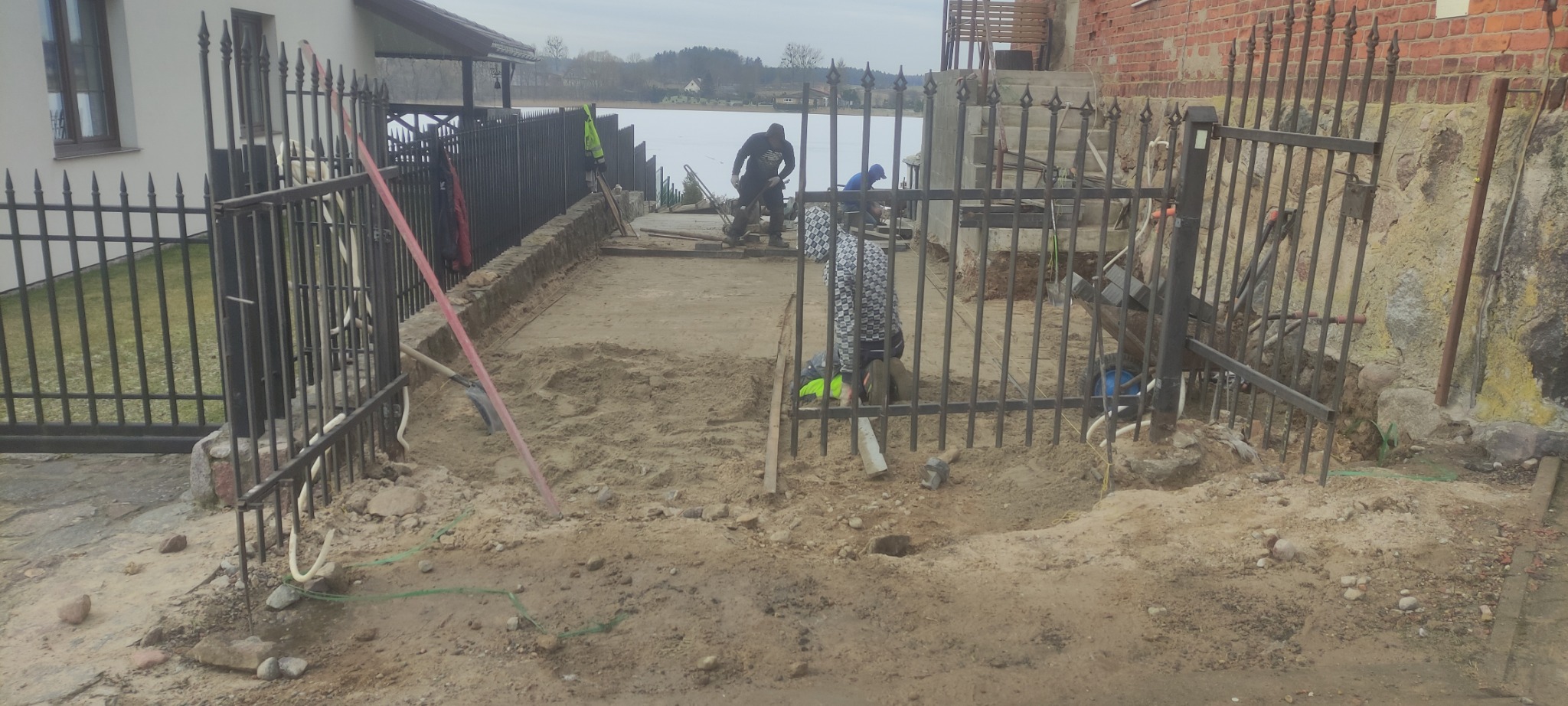 Montaż metalowego ogrodzenia z ozdobnymi grotami na tle jeziora, widoczne narzędzia i prace ziemne w toku.