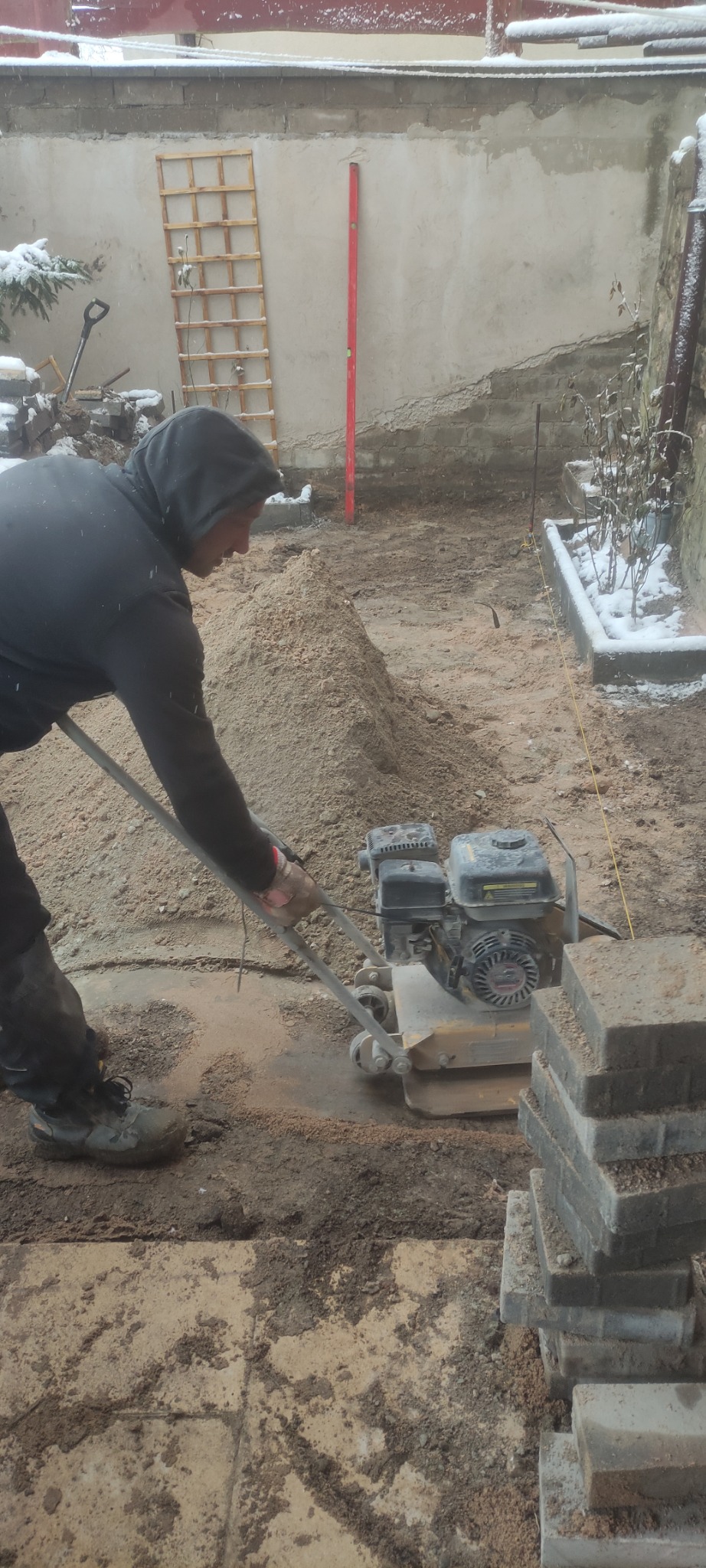 Pracownik w bluzie z kapturem zagęszcza piasek pod kostkę brukową ubijarką w trakcie chłodnego, pochmurnego dnia.