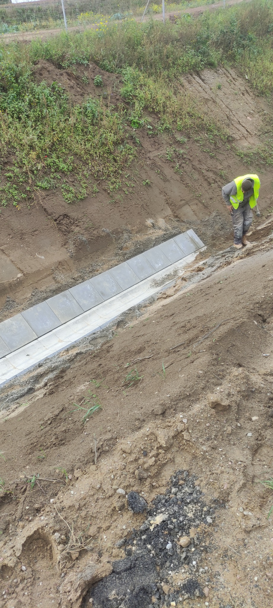 Montaż krawężnika betonowego w rowie przydrożnym, pracownik w odblaskowej kamizelce zagęszcza podłoże ubijakiem, widoczne nierówności terenu i roślinność na skarpie.