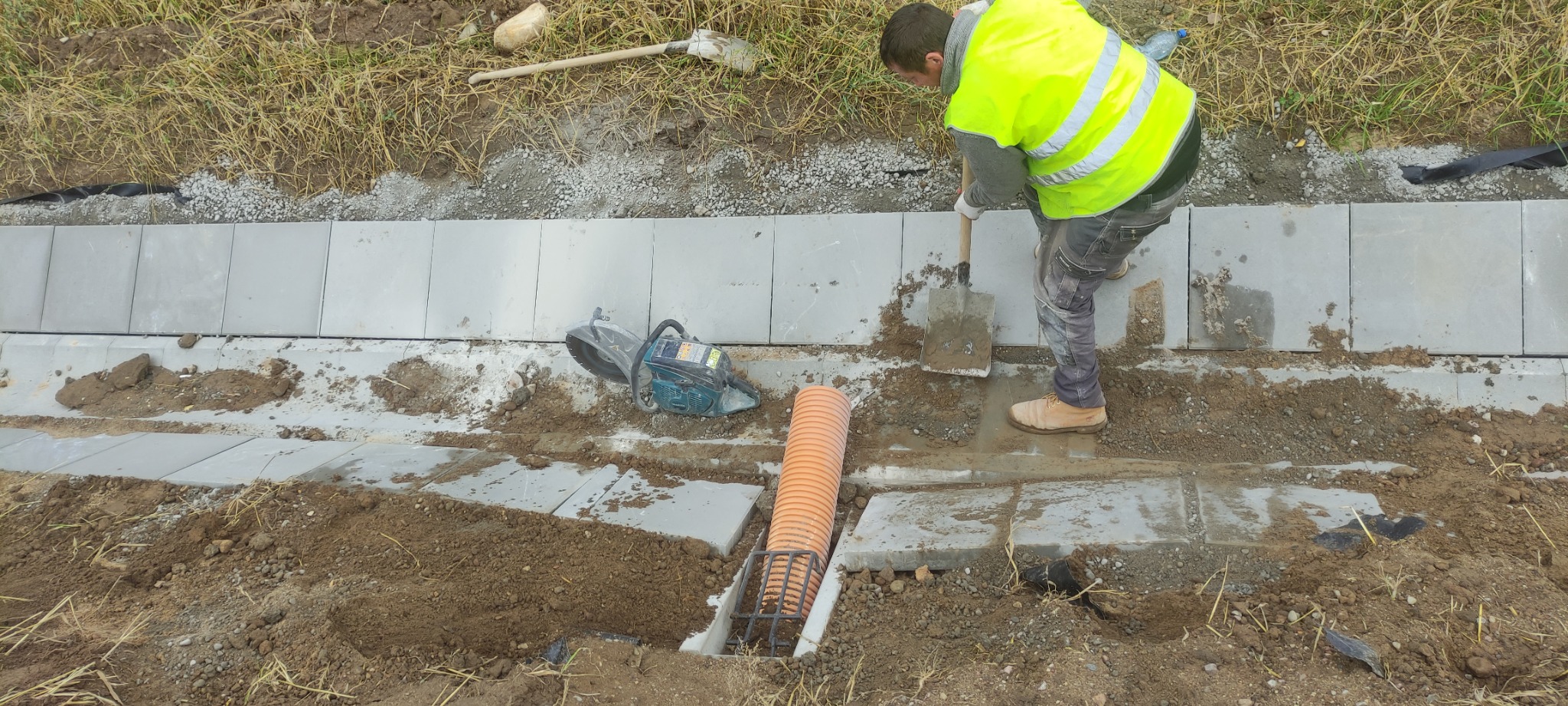 Pracownik w odblaskowej kamizelce wyrównuje podłoże pod obrzeża chodnika przy użyciu szpadla; widoczna piła do cięcia betonu, rura drenarska i ułożone obrzeża.