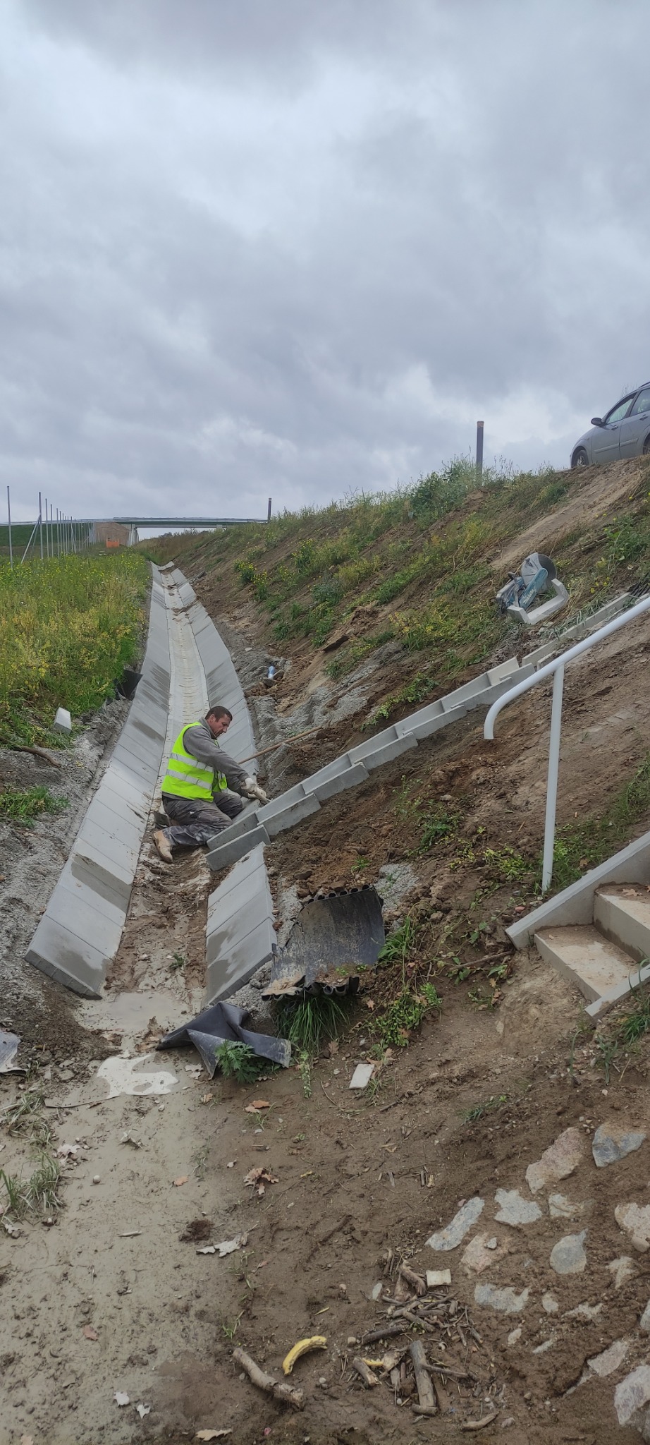 Pracownik w kamizelce odblaskowej układa betonowe elementy koryta odwadniającego w wykopie, widoczne schody z balustradą i samochód w tle.