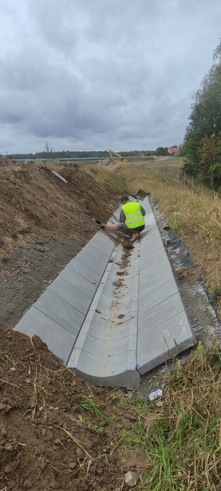 Montaż betonowego koryta odwadniającego na placu budowy, pracownik w żółtej kamizelce poziomuje elementy, w tle koparka i pochmurne niebo.