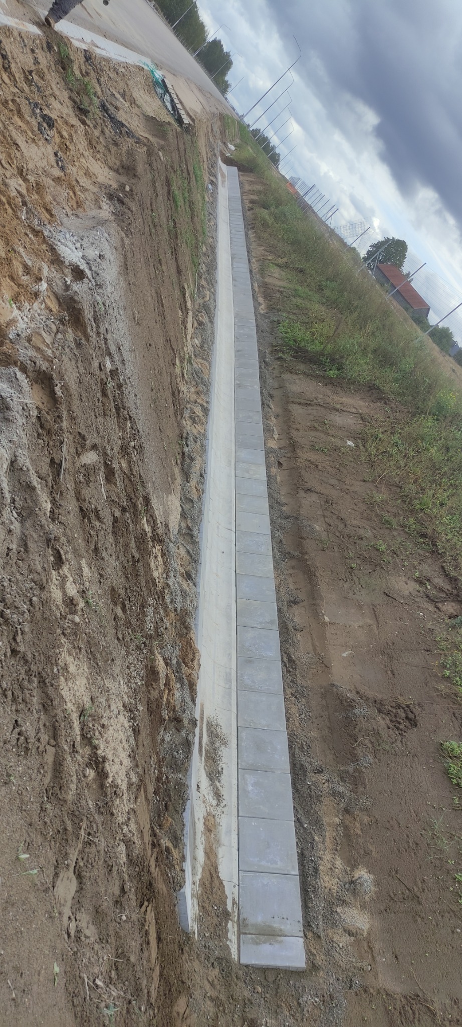 Długi, wąski rów wyłożony szarą kostką brukową wzdłuż drogi, widok z góry, pochmurne niebo w tle.