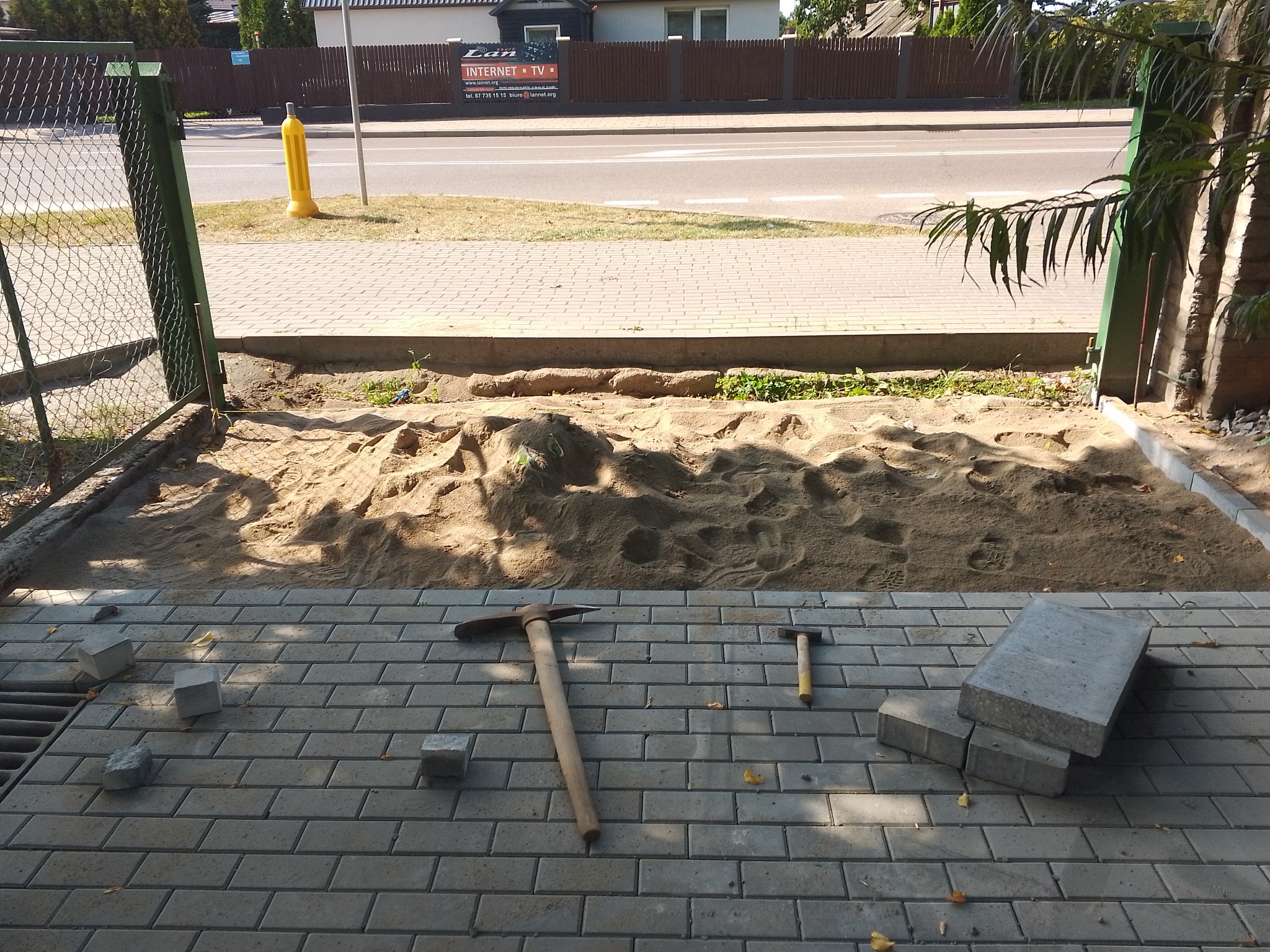 Przygotowanie podłoża pod chodnik z kostki brukowej: wyrównany piasek, porozrzucane kostki, czekany i fragment gotowego chodnika przy ulicy.