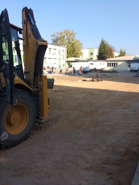 Żółta koparko-ładowarka z widocznymi śladami opon na piasku, w tle grupa robotników w pomarańczowych kombinezonach przygotowuje teren pod budowę, budynki i samochody dostawcze w tle.