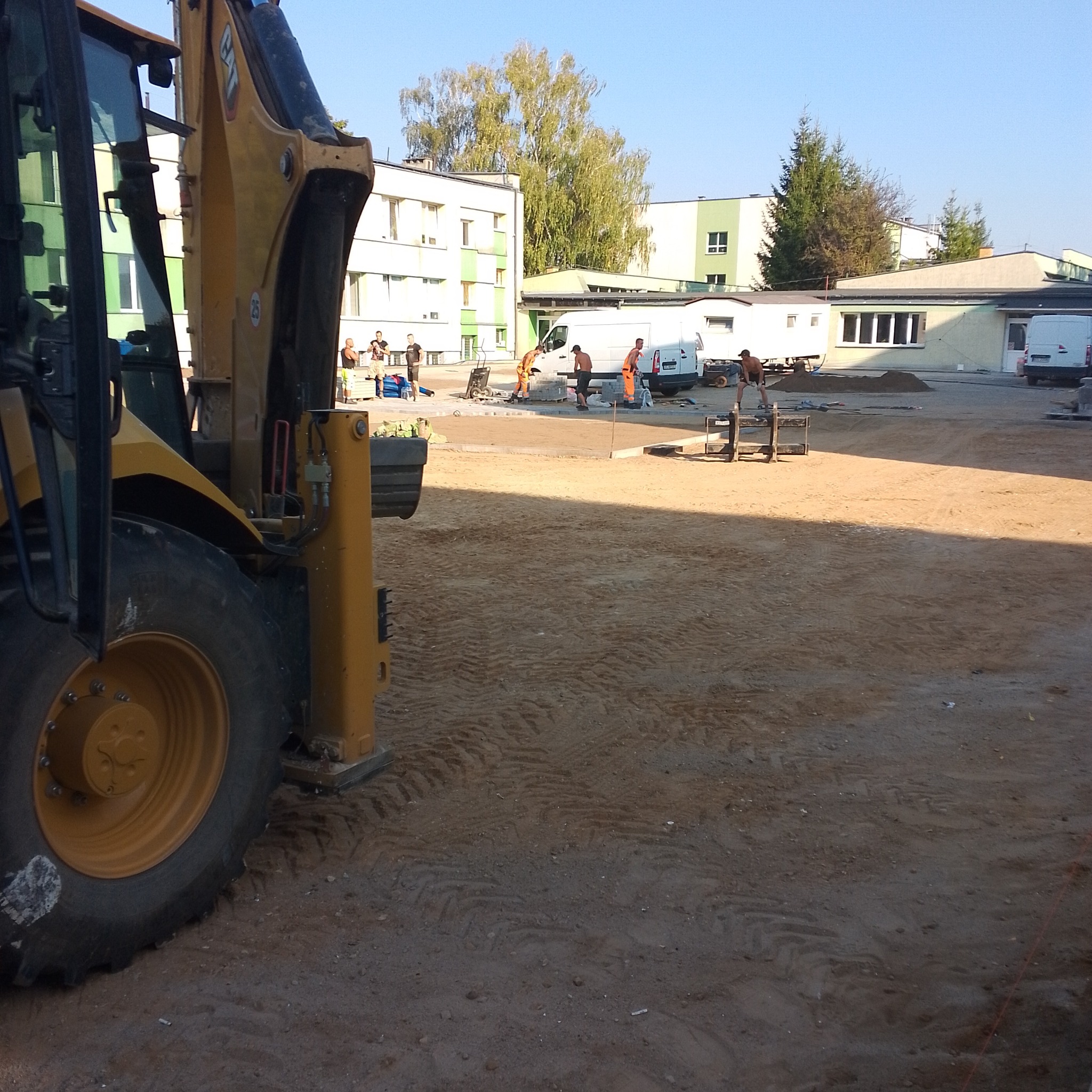 Żółta koparko-ładowarka na placu budowy z pracownikami układającymi nawierzchnię, widoczne budynki w tle i białe busy.