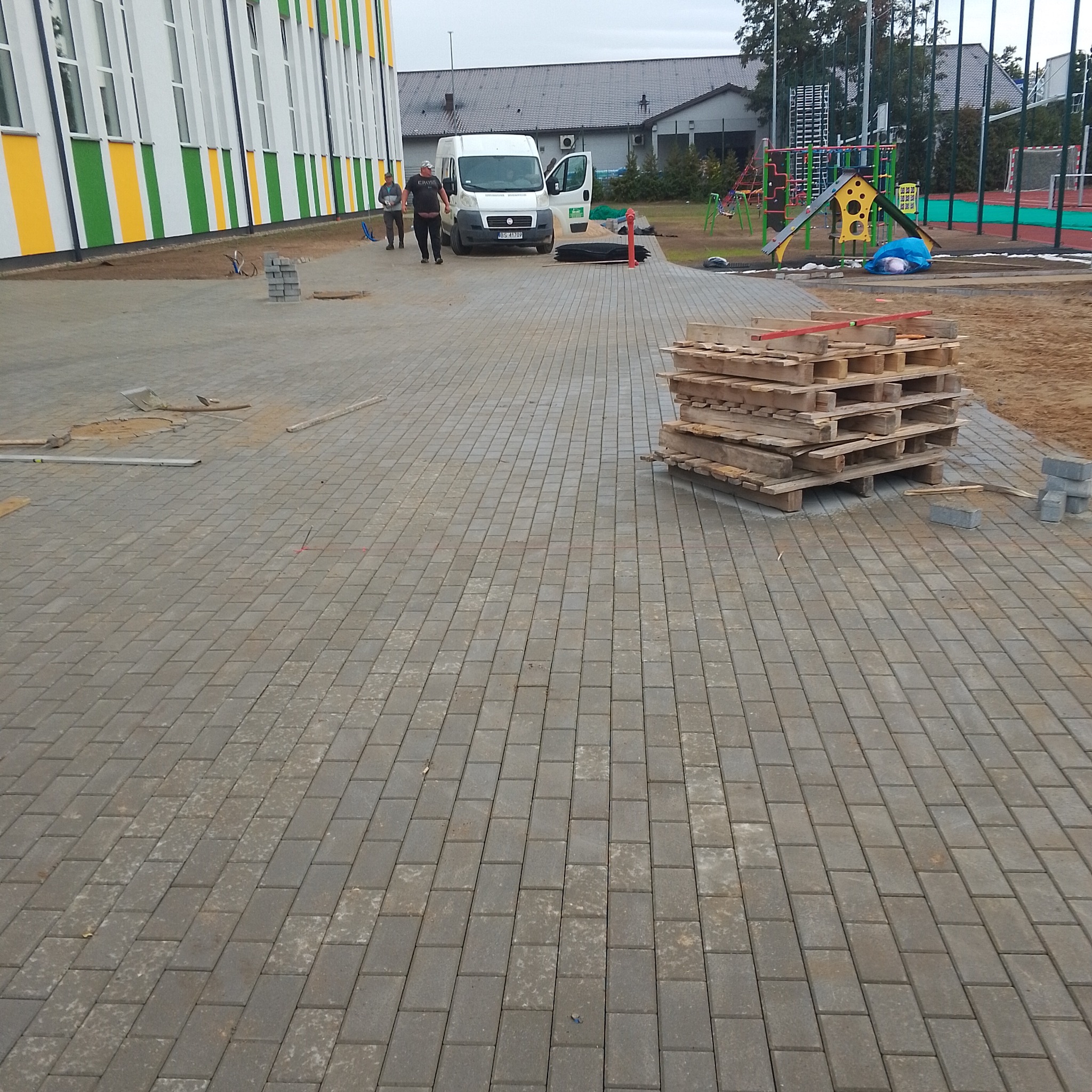 Nowa nawierzchnia z szarej kostki brukowej na placu zabaw szkolnym, paleta z kostką, biały bus dostawczy i dwóch robotników w tle.