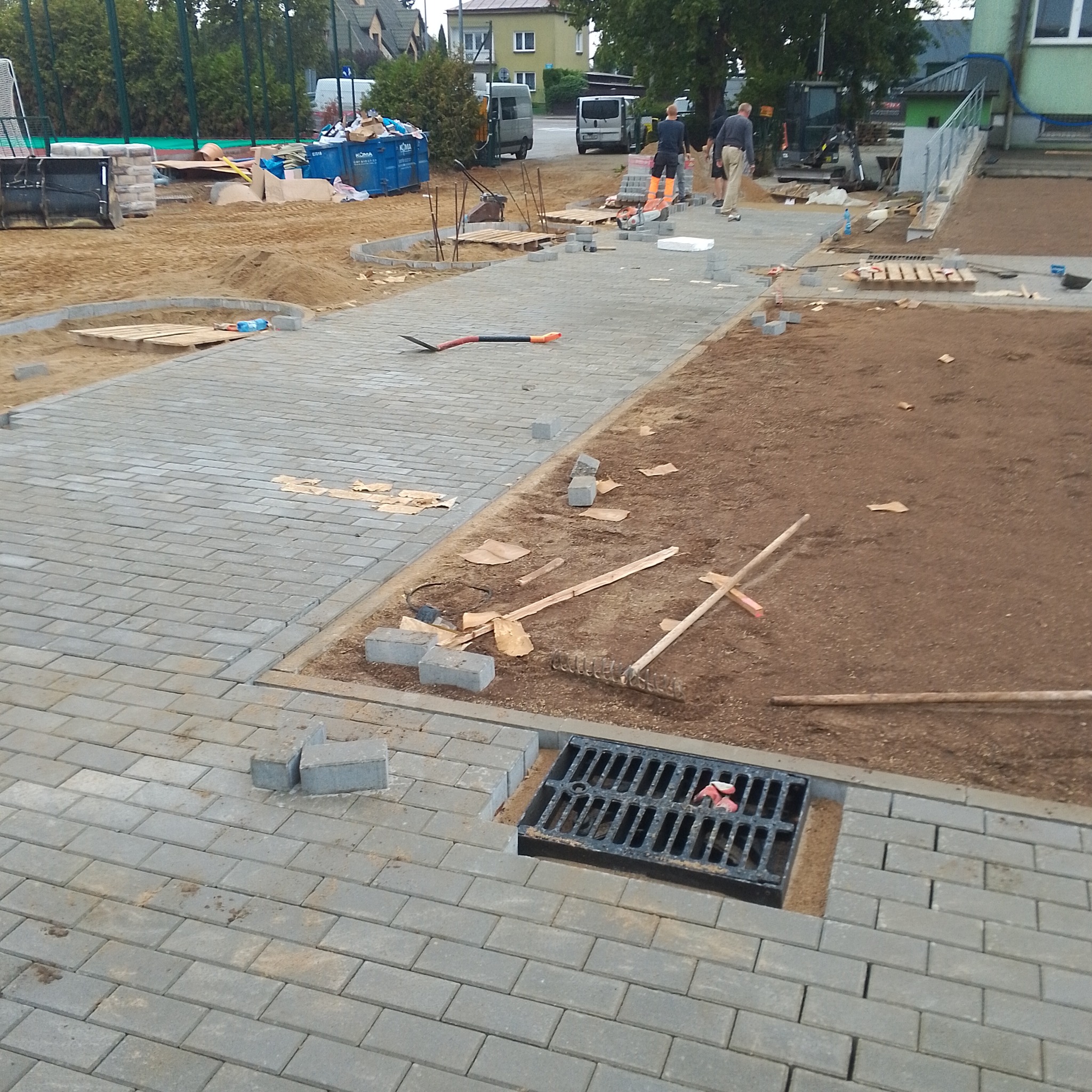Trwa układanie szarej kostki brukowej na nowym chodniku, widoczne narzędzia, piasek i niezakończone fragmenty nawierzchni, w tle budynki i pracownicy.