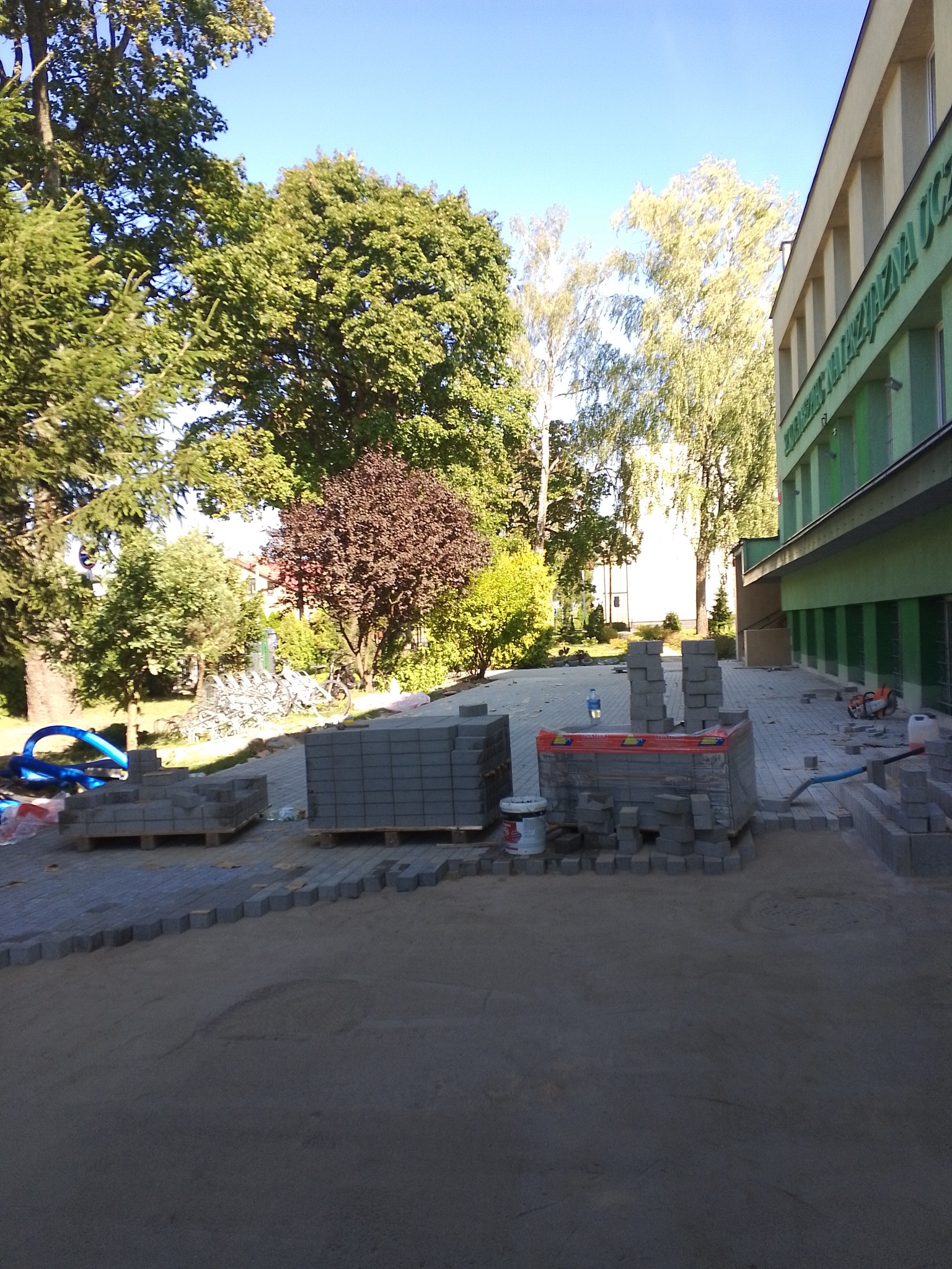 Układanie szarej kostki brukowej przed budynkiem szkoły w słoneczny dzień, widoczne palety z kostką, piasek i częściowo ułożona nawierzchnia.