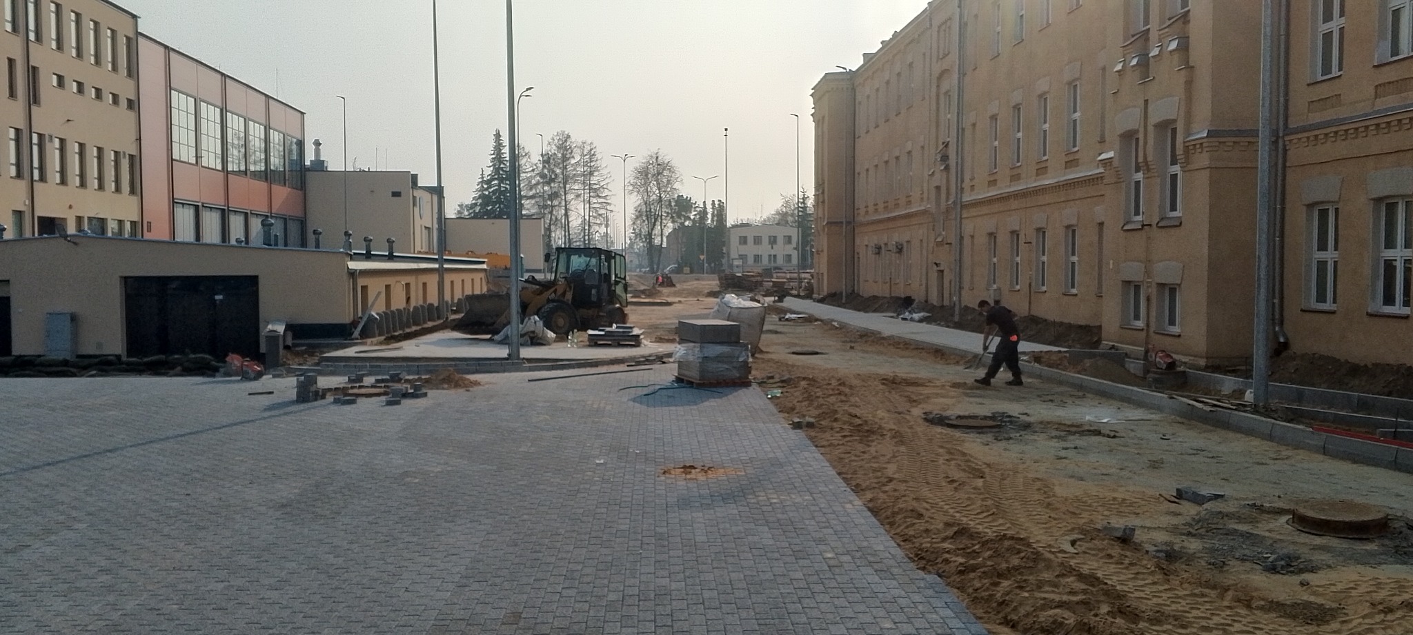 Rozległy teren z częściowo ułożoną szarą kostką brukową, koparka i pracownik wyrównujący piasek pod dalszą część nawierzchni, budynek w tle.