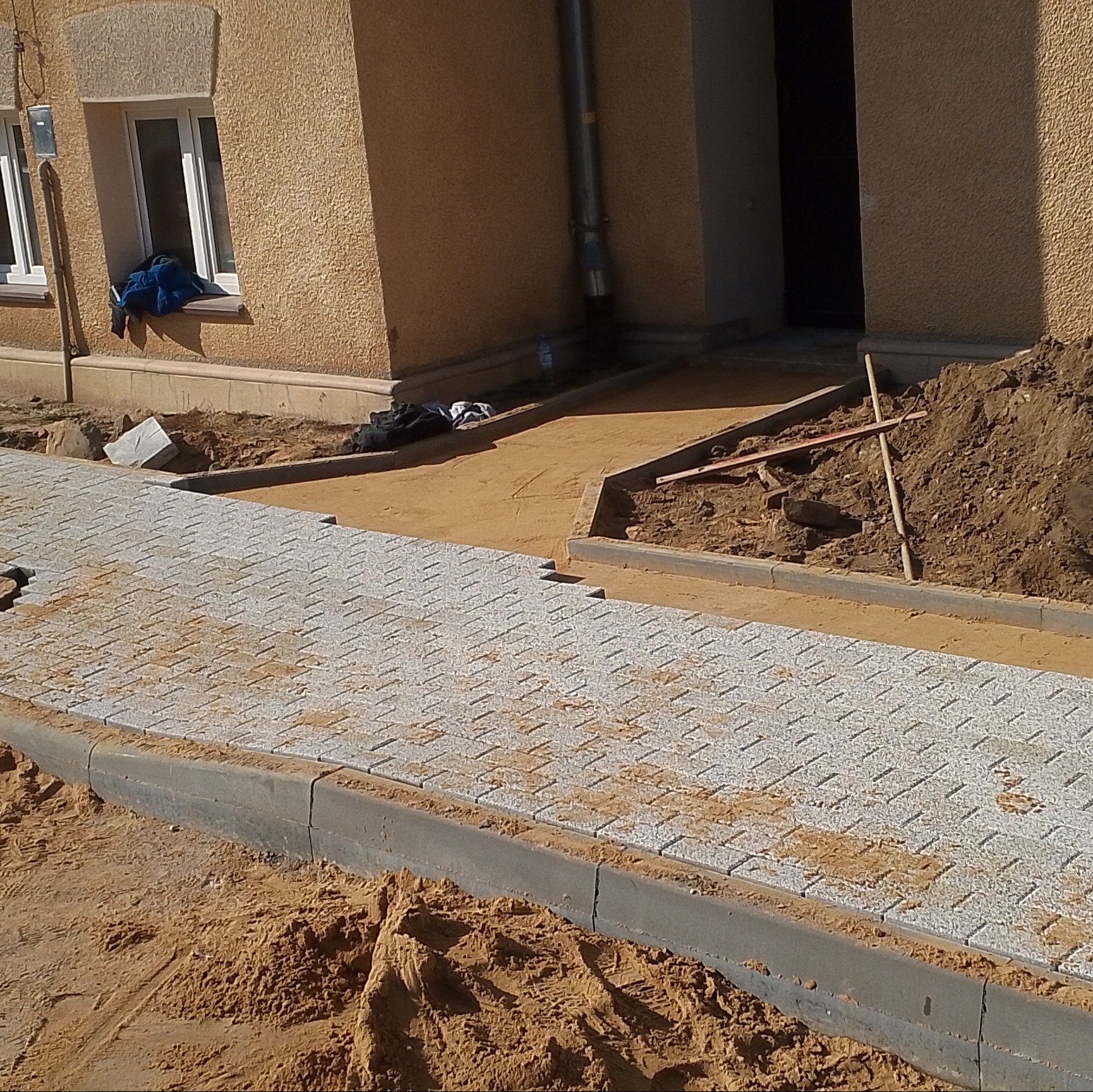 Fragment nowo układanej ścieżki z jasnoszarej kostki brukowej o wzorze jodełki, obrzeża z szarego betonu, piasek między kostkami, budynek z oknem w tle, widoczne narzędzia i pryzma ziemi.
