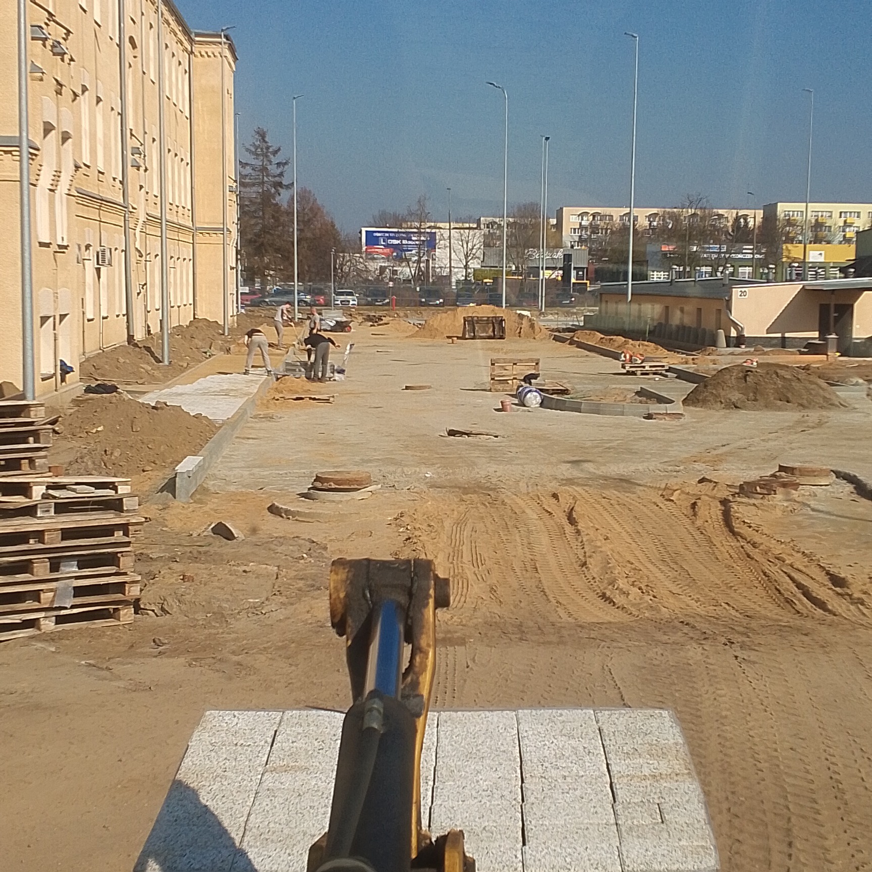 Widok z perspektywy operatora maszyny układającej jasnoszarą kostkę granitową na placu budowy, w tle ekipa robotników, budynek i uliczne latarnie.