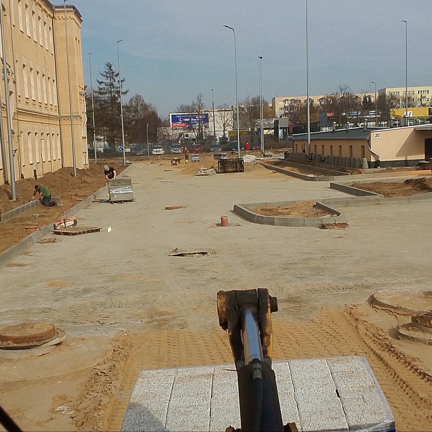 Plac budowy z widocznymi pracami przygotowawczymi pod ułożenie nawierzchni, w tle budynki i pracownicy, na pierwszym planie maszyna z paletą kostki.