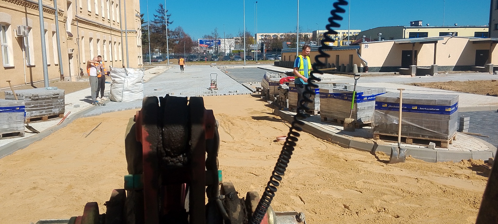 Widok z perspektywy operatora maszyny do układania kostki brukowej na placu budowy, z pracownikami i paletami kostki w tle.