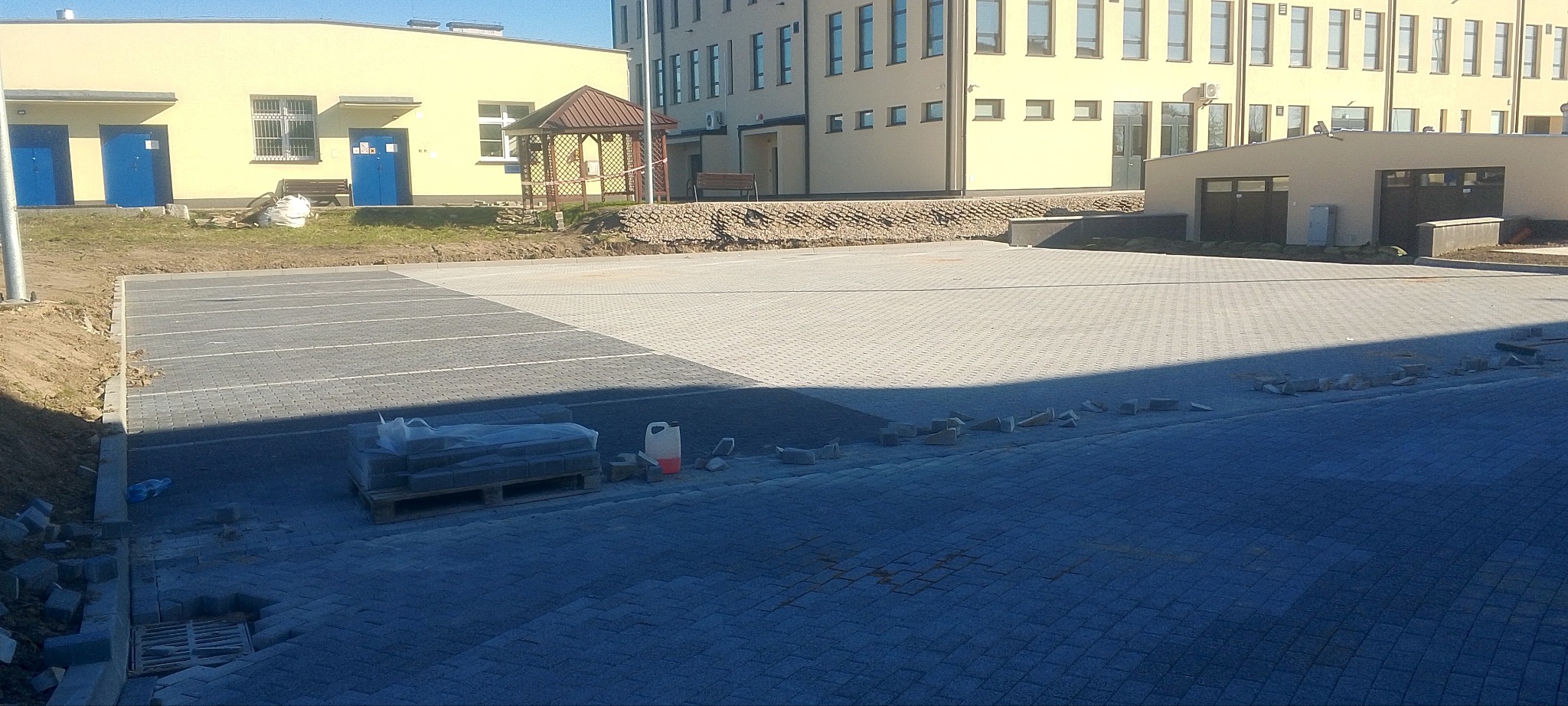 Niedokończona nawierzchnia parkingu z kostki brukowej w odcieniach szarości, paleta z zapasem kostki, widoczne budynki w tle.