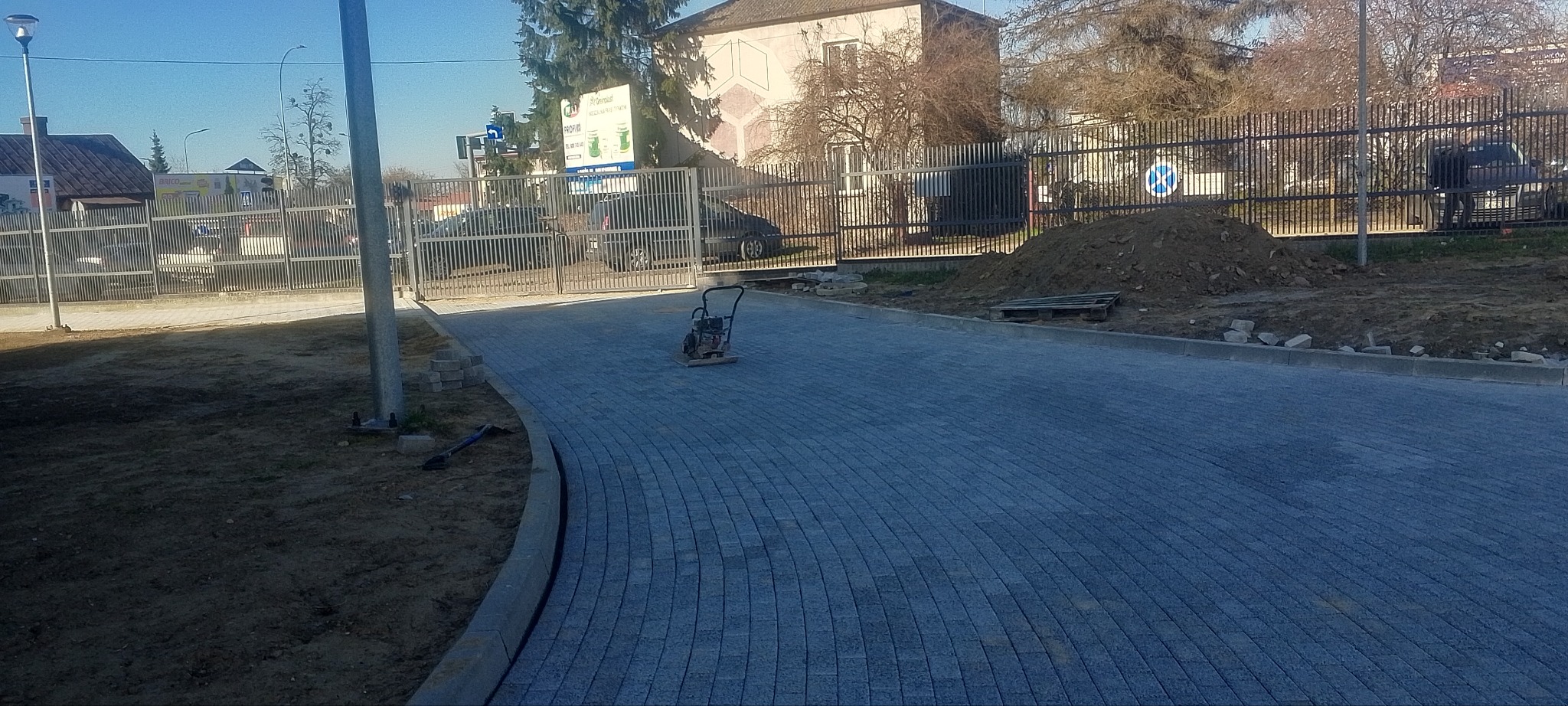 Świeżo ułożona, szara kostka brukowa na zakrzywionym chodniku, zagęszczarka wibracyjna stoi na środku, krawężnik oddziela kostkę od ziemi, w tle brama i budynek gospodarczy.