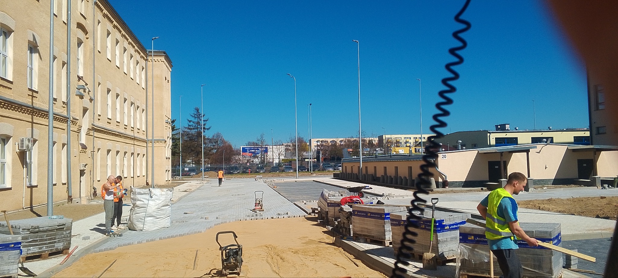 Pracownicy w odblaskowych kamizelkach układają szarą kostkę brukową na placu przed budynkiem, z użyciem ubijarki wibracyjnej i palet z kostką, widok na budynek w tle.