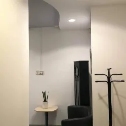 Wnętrze korytarza z kremowymi ścianami, czarnym wieszakiem na ubrania, głośnikiem, małym stolikiem z rośliną i drzwiami z przeszkleniem.