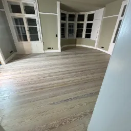 Jasne, odnowione deski podłogowe w pokoju z oknami w białych ramach, widok zza otwartych drzwi.