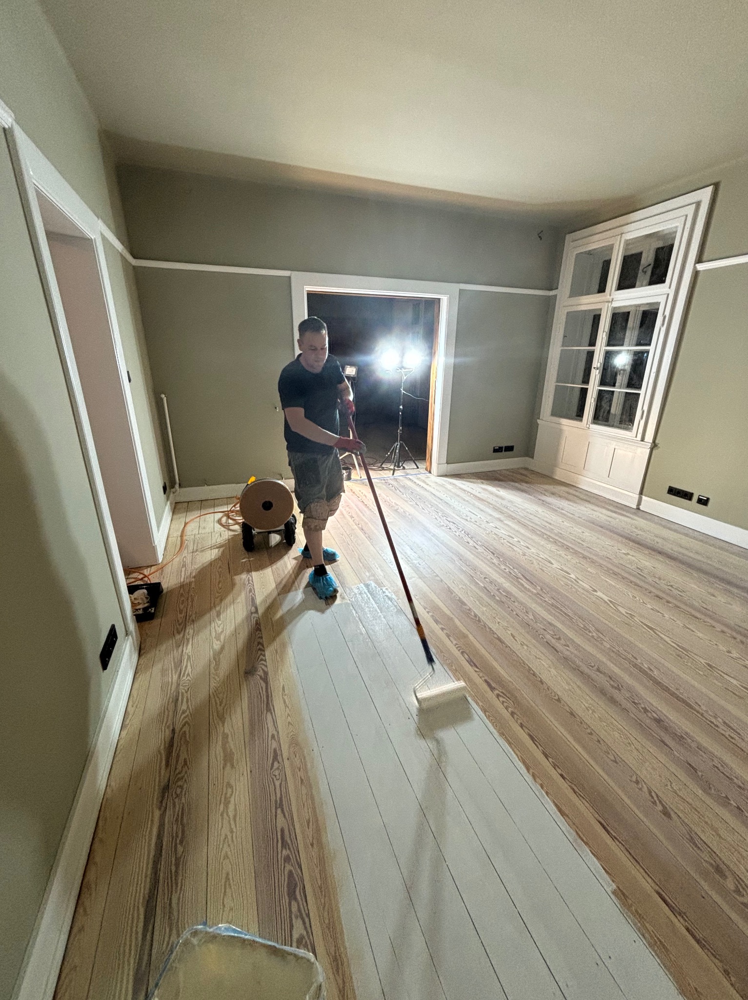 Mężczyzna w niebieskich ochraniaczach na buty maluje drewnianą podłogę wałkiem w przestronnym pokoju z białymi framugami drzwi i okien, widoczny przewód od maszyny do cyklinowania.