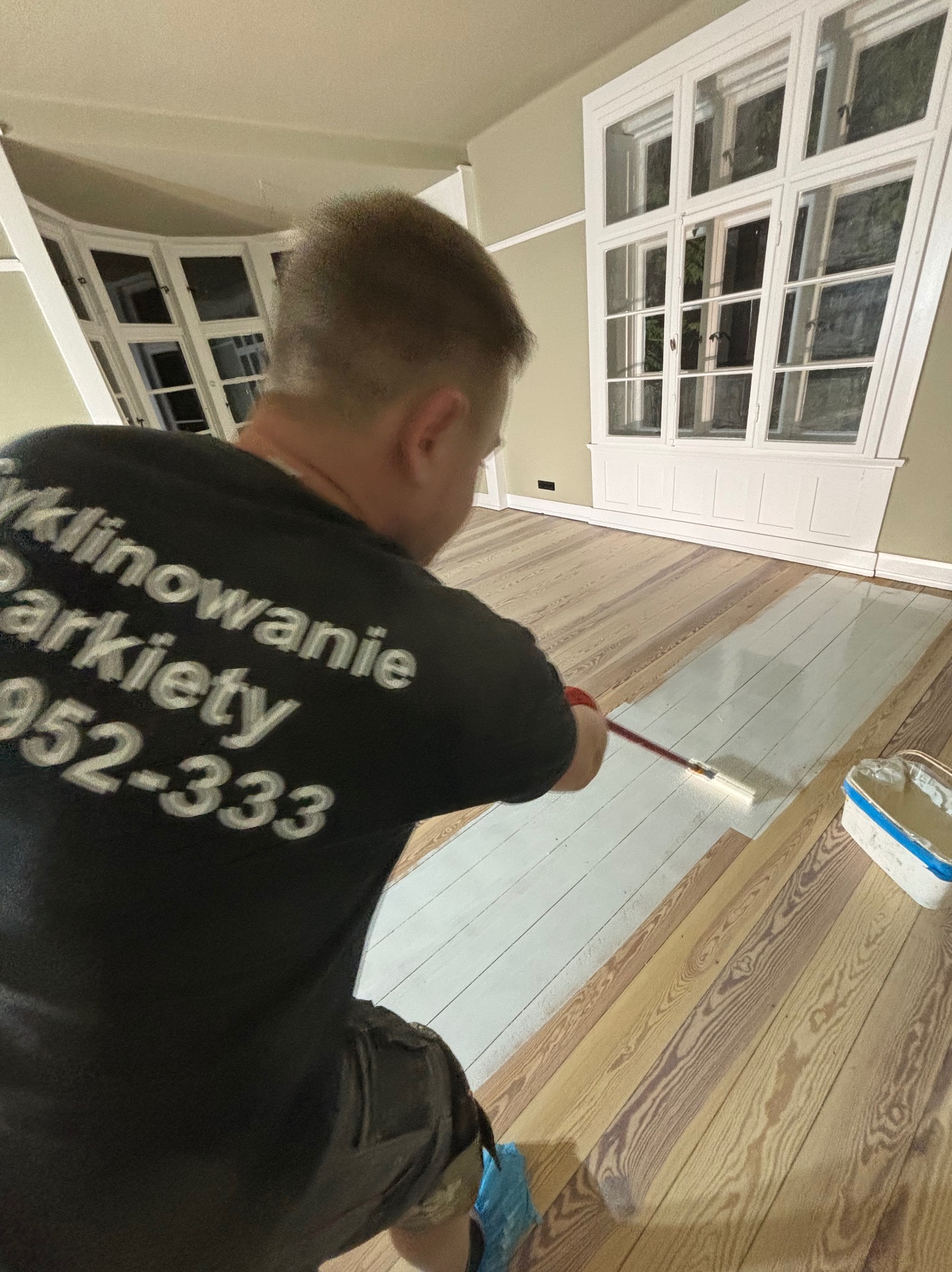 Pracownik w czarnej koszulce z napisem 'Cyklinowanie Parkiety' maluje wałkiem na długim kiju fragment drewnianej podłogi na biało; widoczne duże okna w tle.