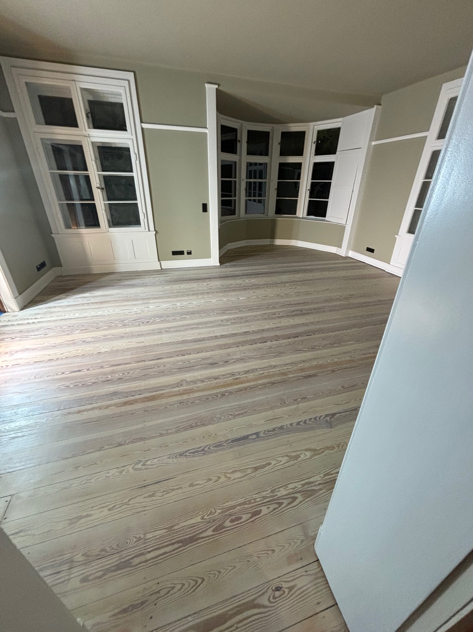 Jasne, odnowione deski podłogowe w pokoju z oknami w białych ramach, widok zza otwartych drzwi.