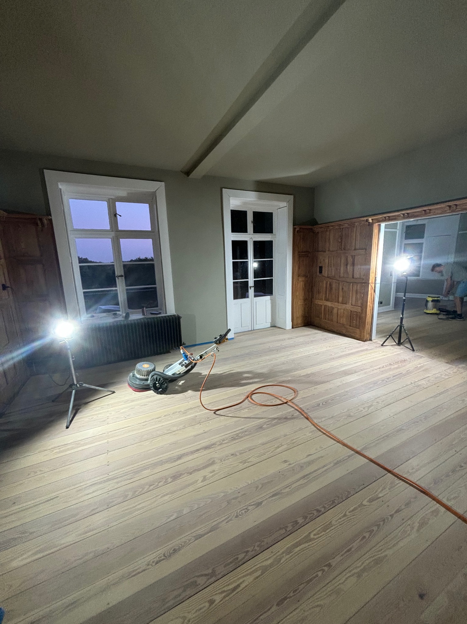 Pomieszczenie z drewnianą podłogą w trakcie cyklinowania; widoczna cykliniarka, lampa oświetleniowa na statywie oraz częściowo wycyklinowana, jasna podłoga z widocznym rysunkiem drewna.