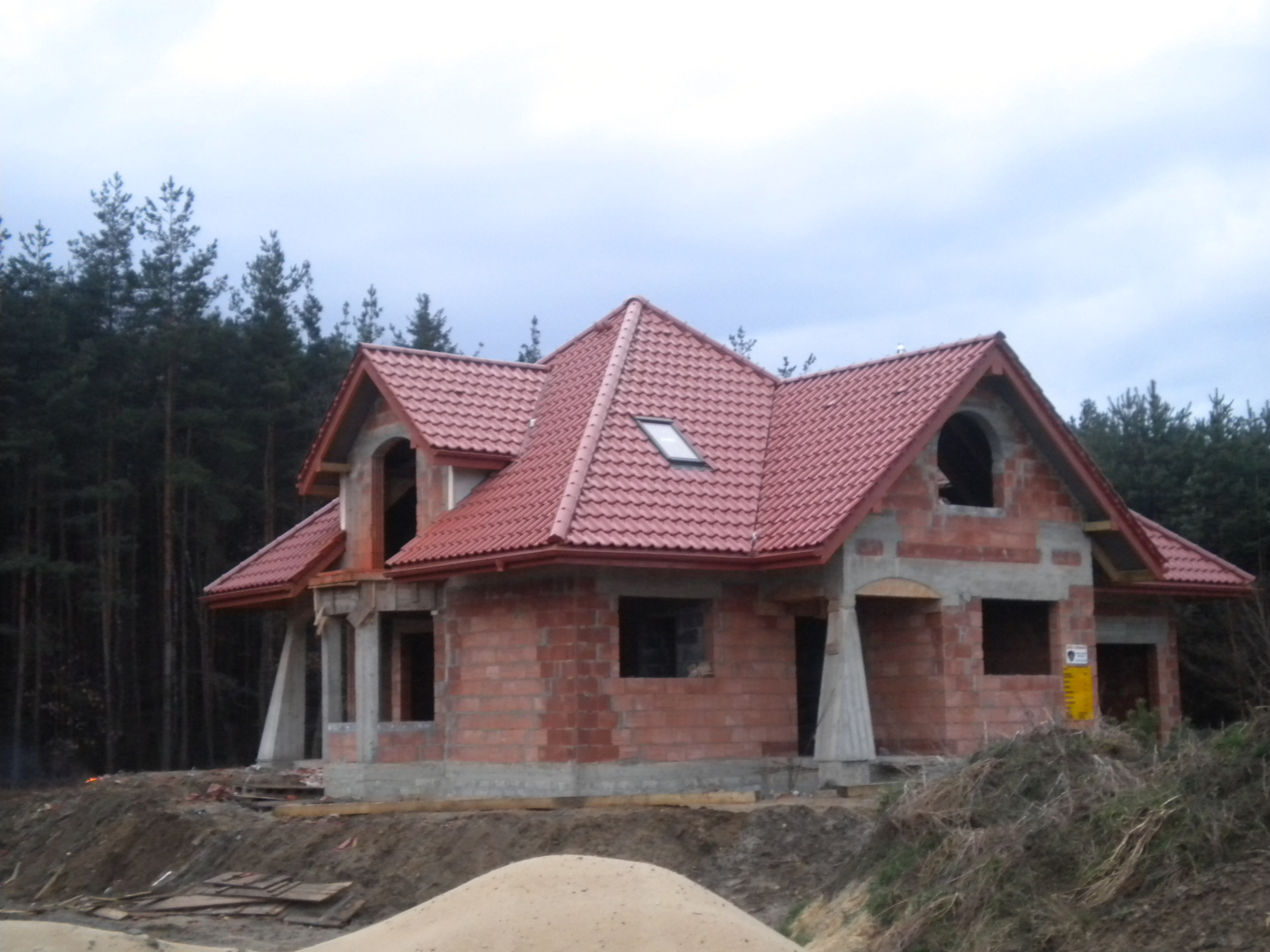 Dom w stanie surowym zamkniętym z czerwoną dachówką, widoczne okno dachowe i orynnowanie, na tle lasu.