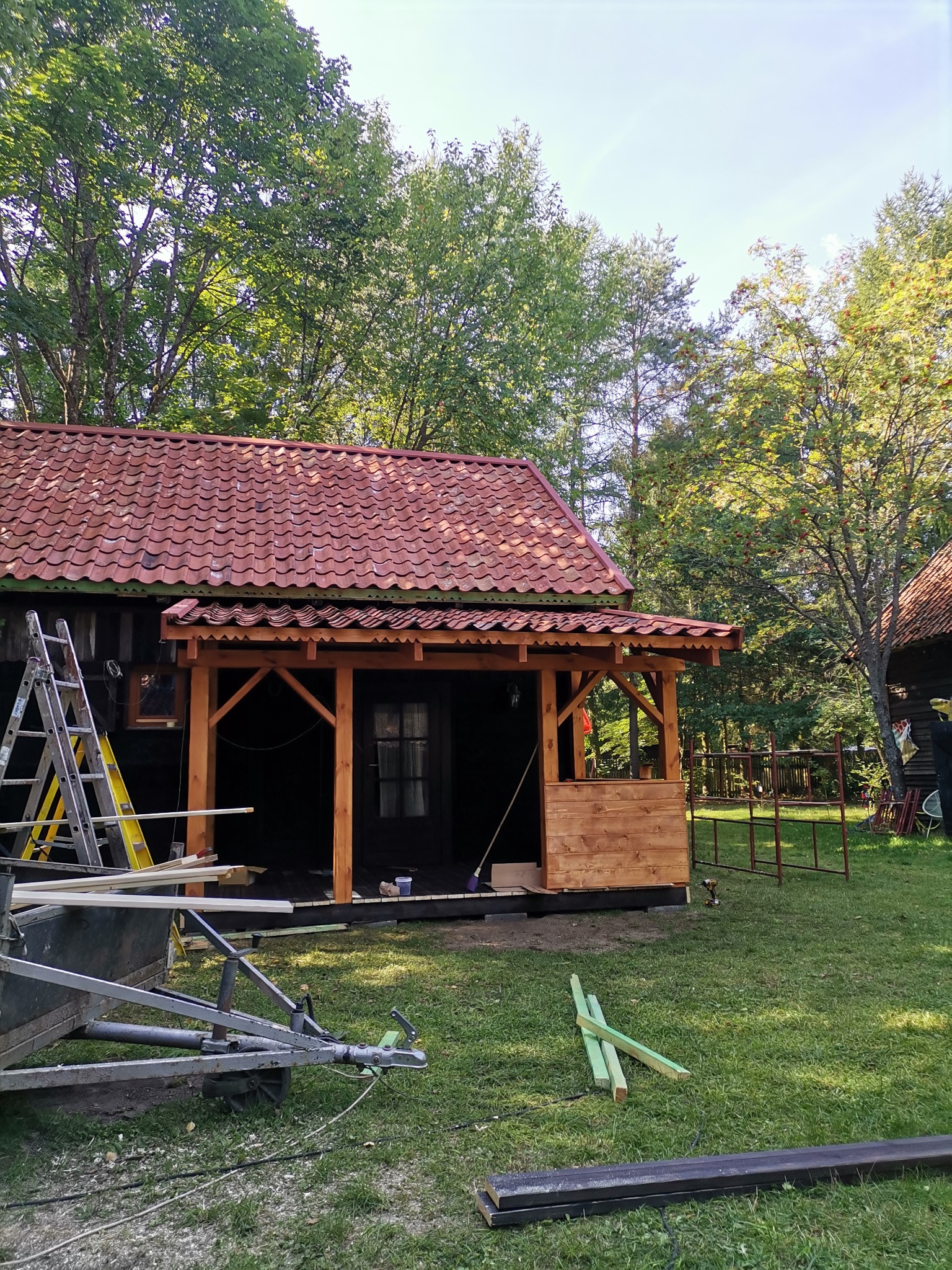 Dom z ciemnego drewna z nową, jasną, drewnianą werandą w trakcie budowy, widoczne narzędzia i materiały budowlane na trawniku.