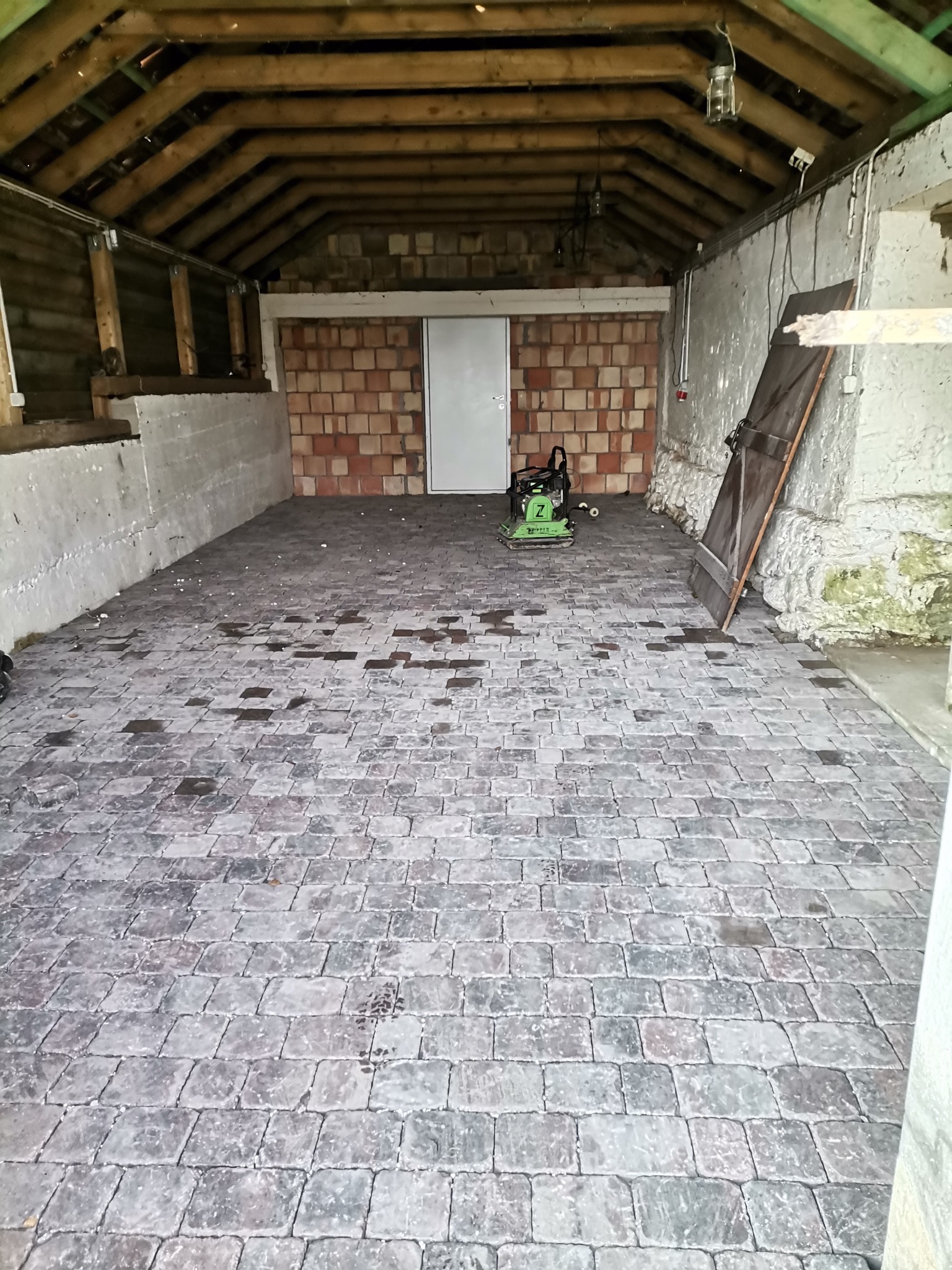 Układanie kostki brukowej wewnątrz pomieszczenia, widoczny ubijak wibracyjny i fragmenty ceglanej ściany oraz drewniany strop.