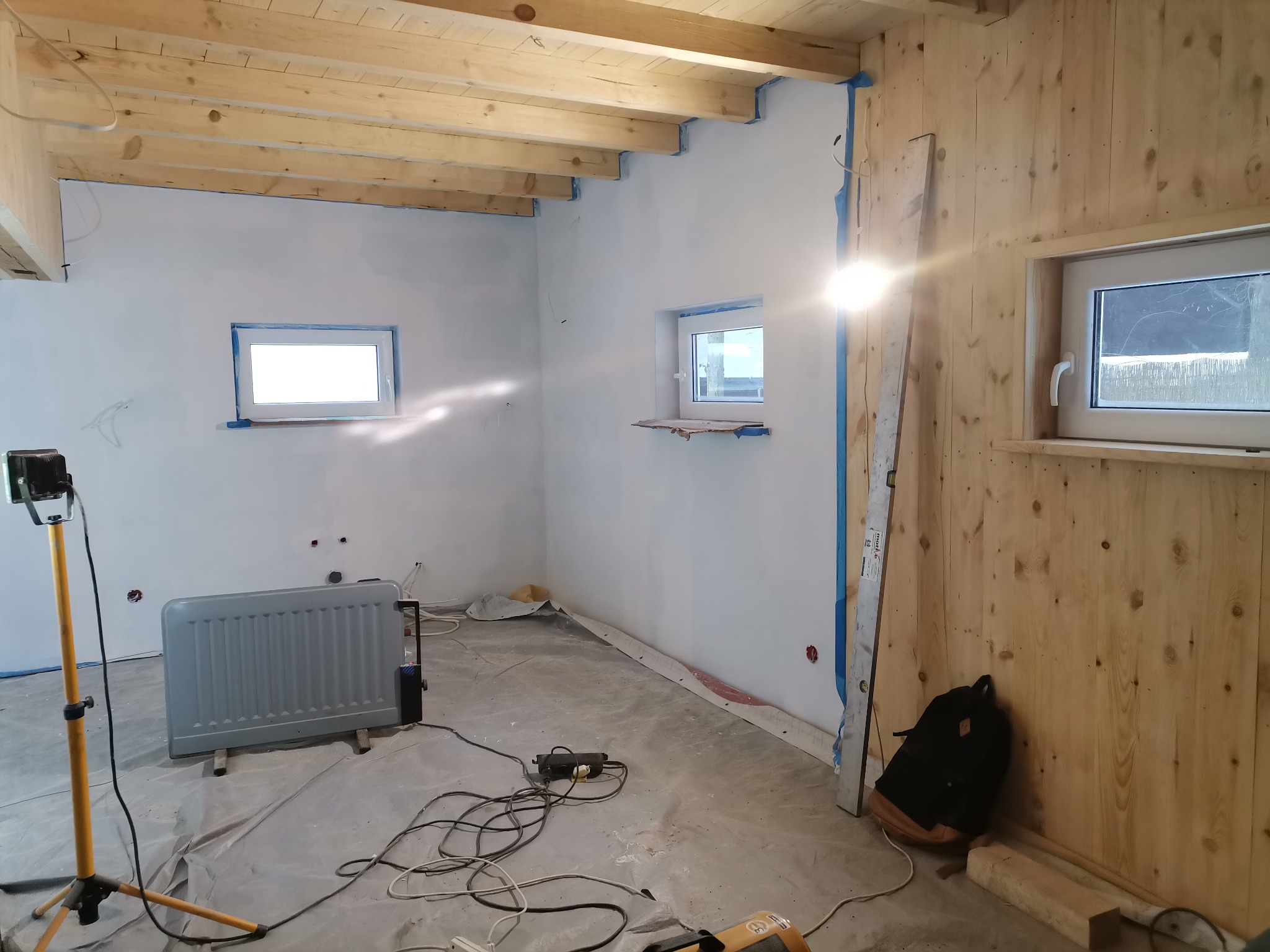 Wnętrze pokoju w trakcie prac wykończeniowych: drewniana boazeria na jednej ścianie, dwie ściany pomalowane na biało, drewniany sufit z widocznymi belkami, okna zabezpieczone taśmą malarską,...