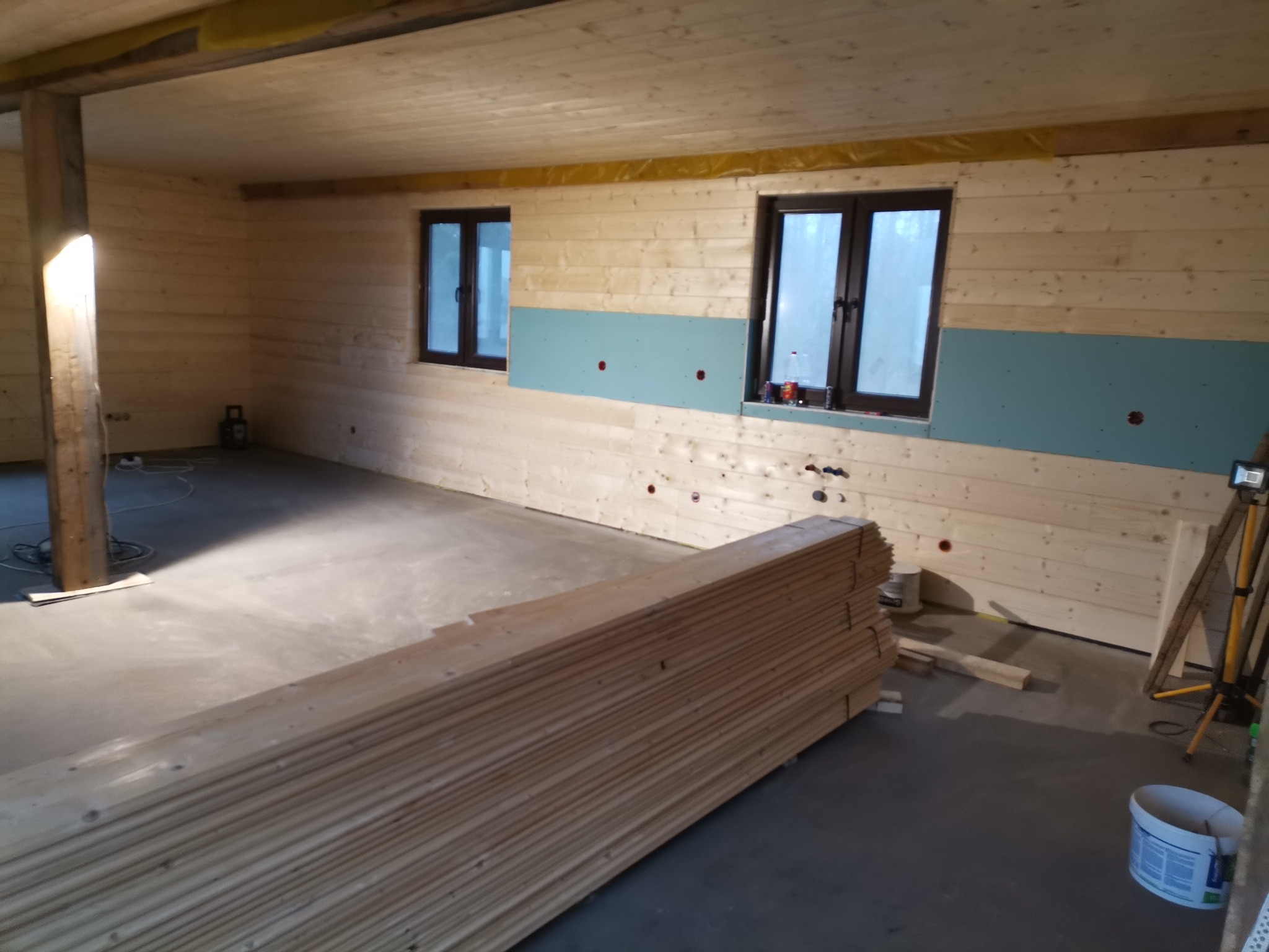 Wnętrze w trakcie wykańczania: ściany i sufit obłożone boazerią, częściowo zamontowane płyty gipsowo-kartonowe, ułożone deski na podłodze, widoczne okna i narzędzia.