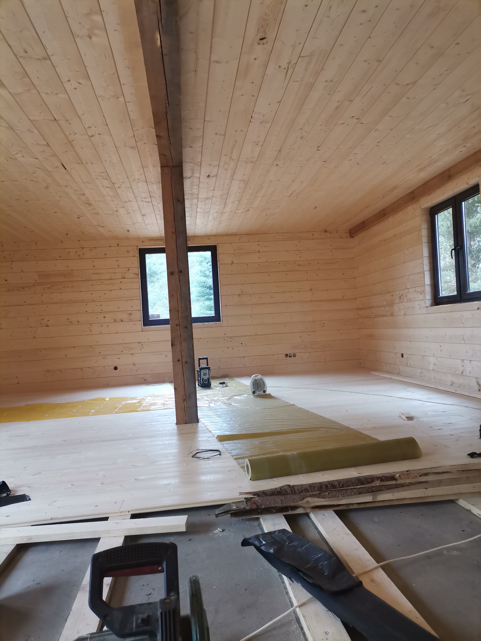 Wnętrze drewnianego domu w trakcie wykończenia, widoczne drewniane ściany i sufit, izolacja podłogowa w rolce, narzędzia budowlane w dolnej części kadru, oraz pionowa belka konstrukcyjna.