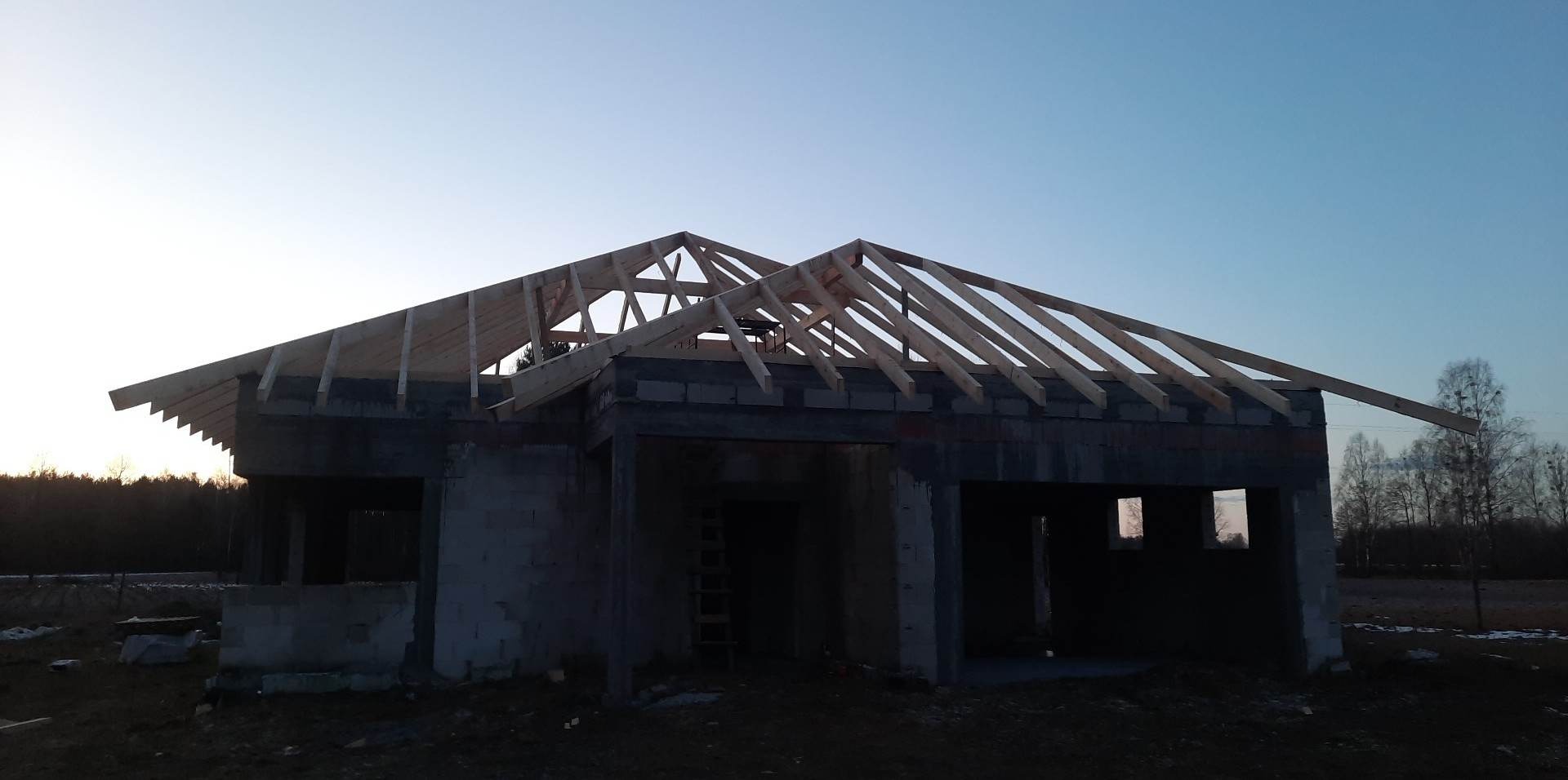 Surowy stan domu jednorodzinnego z widoczną konstrukcją drewnianą dachu na tle zachodzącego słońca.