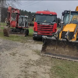 Ciężki sprzęt budowlany: koparka Atlas, czerwona ciężarówka Scania i żółta ładowarka JCB zaparkowane na poboczu drogi wiejskiej przy drzewach.