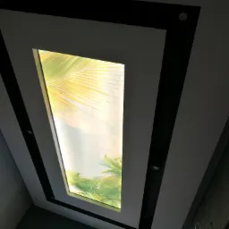 Podwieszany sufit z podświetlanym panelem imitującym okno z widokiem na palmy, czarne listwy dekoracyjne, widoczne niedokończone wykończenie ściany.