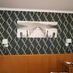 Wzorzysta, ciemna tapeta w romby z białymi obwódkami, ozdobiona czarno-białym obrazem molo, nad brązowym zagłówkiem łóżka i stolikiem nocnym z drobnymi narzędziami w pokoju w Siedlcach.