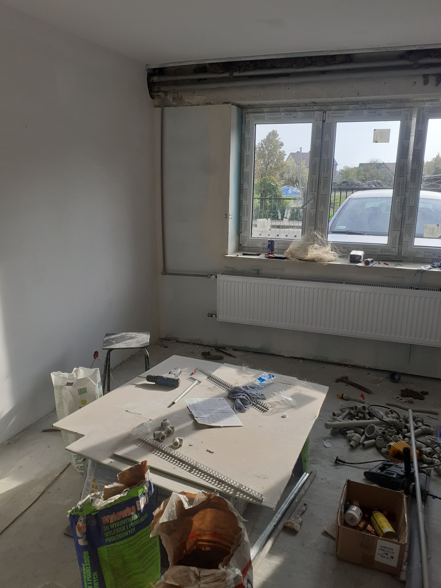 Wnętrze w trakcie remontu: surowy stan, widoczne narzędzia, rury PCV, worki z zaprawą, odsłonięte okno z widokiem na zewnątrz, grzejnik centralnego ogrzewania.