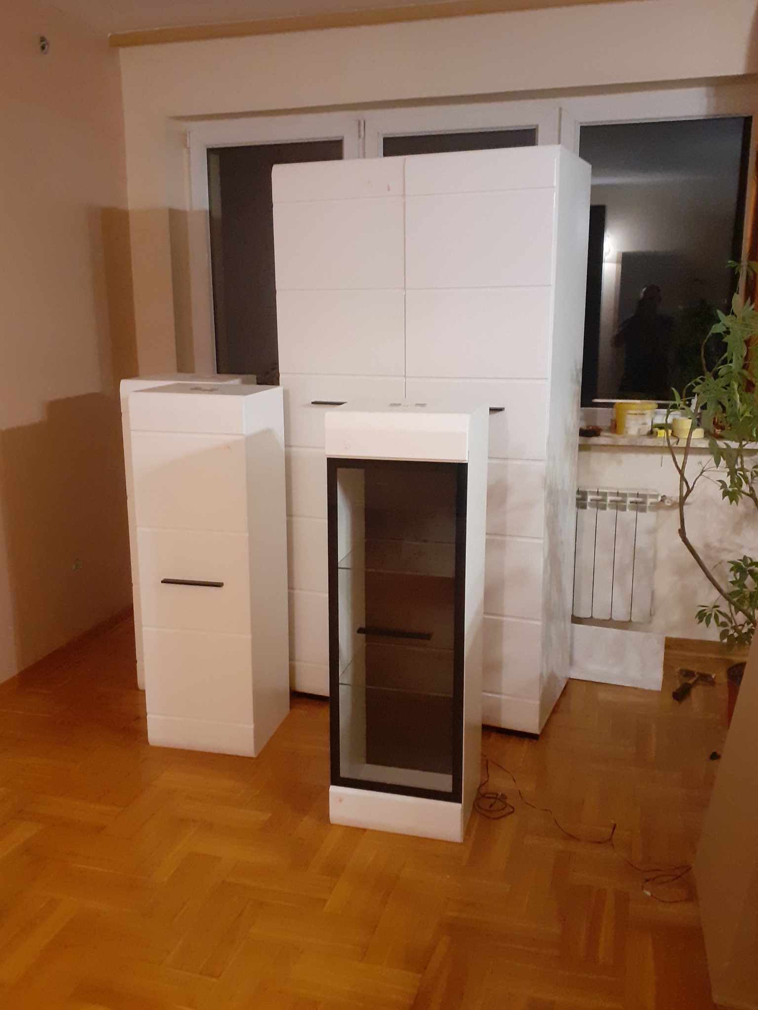 Ustawione w pokoju trzy białe szafki z ciemnymi uchwytami na tle okna i grzejnika, na parkiecie ułożonym w jodełkę.