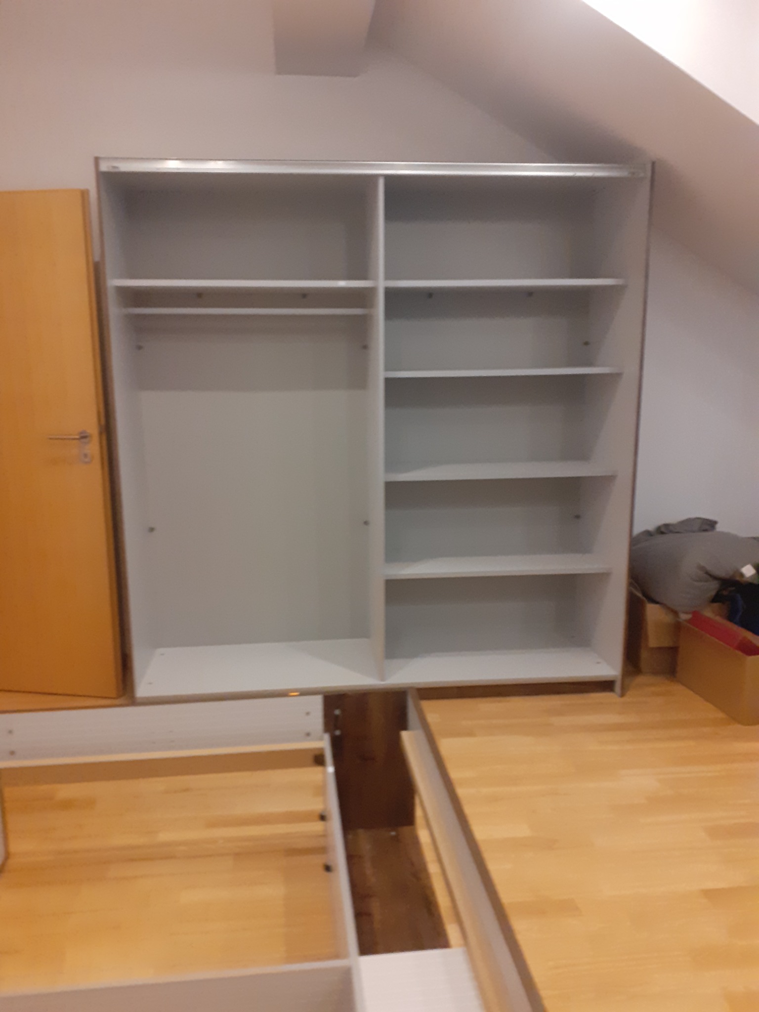 Montaż szafy wnękowej z półkami i drążkiem w pokoju na poddaszu, widoczne elementy łóżka w trakcie składania.