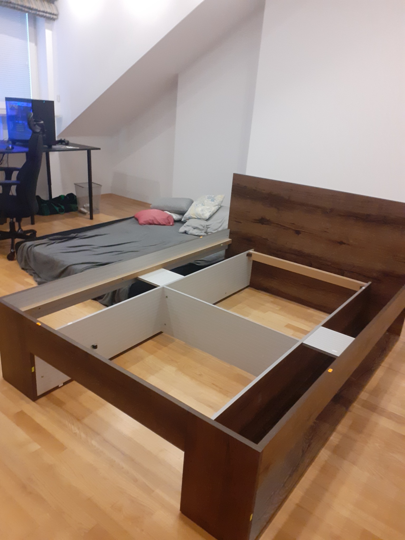 Montaż ramy łóżka z ciemnego drewna z białymi elementami w pokoju z ukośnym sufitem, widoczny materac na podłodze i biurko z komputerem w tle.