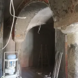 Wnętrze w trakcie remontu z odsłoniętą cegłą w łukach, kolumna z szorstką fakturą, maszyna do mieszania zaprawy, drabina oparta o ścianę, widoczne ślady prac murarskich i instalacyjnych.