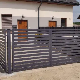 Nowoczesne, poziome ogrodzenie panelowe w kolorze grafitowym przed dwoma domami bliźniaczymi z brązowymi drzwiami i szarą elewacją, widoczne skrzynki na gaz i prąd.