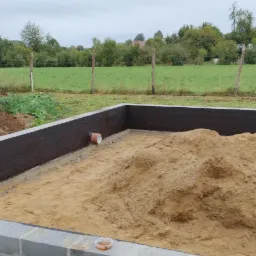 Fundament z izolacją przeciwwilgociową, wypełniony piaskiem, z widocznym wykopem ziemi i koparką w tle, na tle zieleni i zabudowań.