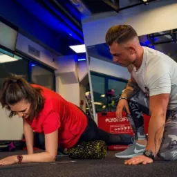 Kobieta w czerwonej koszulce wykonuje ćwiczenie rozciągające z rollerem pod nadzorem mężczyzny w sportowym stroju w siłowni z niebieskim oświetleniem.