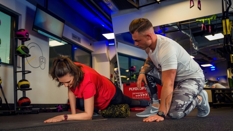 Kobieta w czerwonej koszulce wykonuje ćwiczenie rozciągające z rollerem pod nadzorem mężczyzny w sportowym stroju w siłowni z niebieskim oświetleniem.