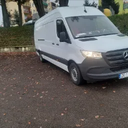 Biały samochód dostawczy Mercedes-Benz Sprinter zaparkowany na parkingu pokrytym opadłymi liśćmi, z widoczną tablicą rejestracyjną ZK 1180H i budynkiem w tle.